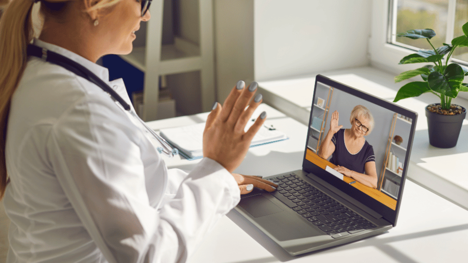 A doctor on a virtual telehealth call waving to the elderly female patient on the screen.